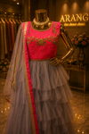 A pink and gray tiered tulle ethnic gown displayed on a gold mannequin in a boutique. The dress features a bright pink embroidered bodice, a layered gray tulle skirt, and a matching pink and gray dupatta with gold borders.