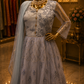 A formal Anarkali gown displayed on a mannequin in a boutique setting. The dress is a dusty lavender-gray color with intricate silver-blue floral embroidery, a gold buttoned neckline, sheer long sleeves, and a matching sheer dupatta draped over one shoulder.