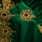 A close-up of a rich emerald green fabric, likely a saree or traditional garment, featuring intricate gold floral embroidery and small red and green stone accents along the border.